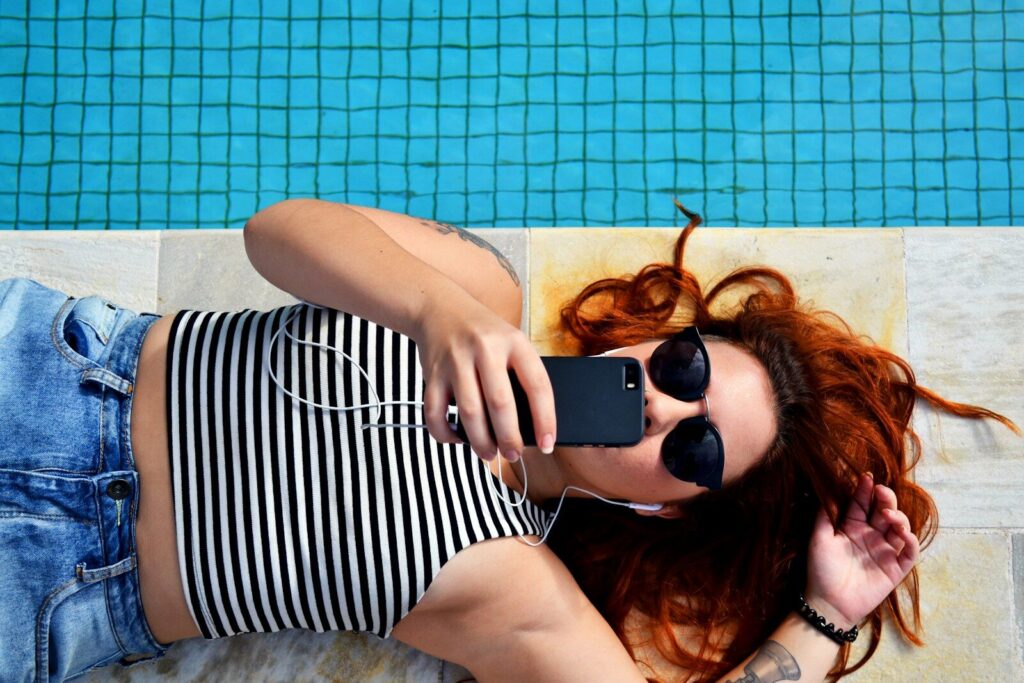 Kvinde ligger ned ved siden af en pool og kigger på sin mobiltelefon